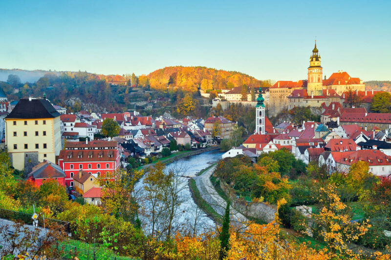 Vista panorámica de Cesky Krumlov al atardecer, donde se puede ver el río que atraviesa la ciudad y las casas de tejados anaranjados y su imponente castillo entre las colinas. El paisaje es de un atardecer. Cesky Krumlov es una de las ciudades cerca de Praga para visitar en una escapada.